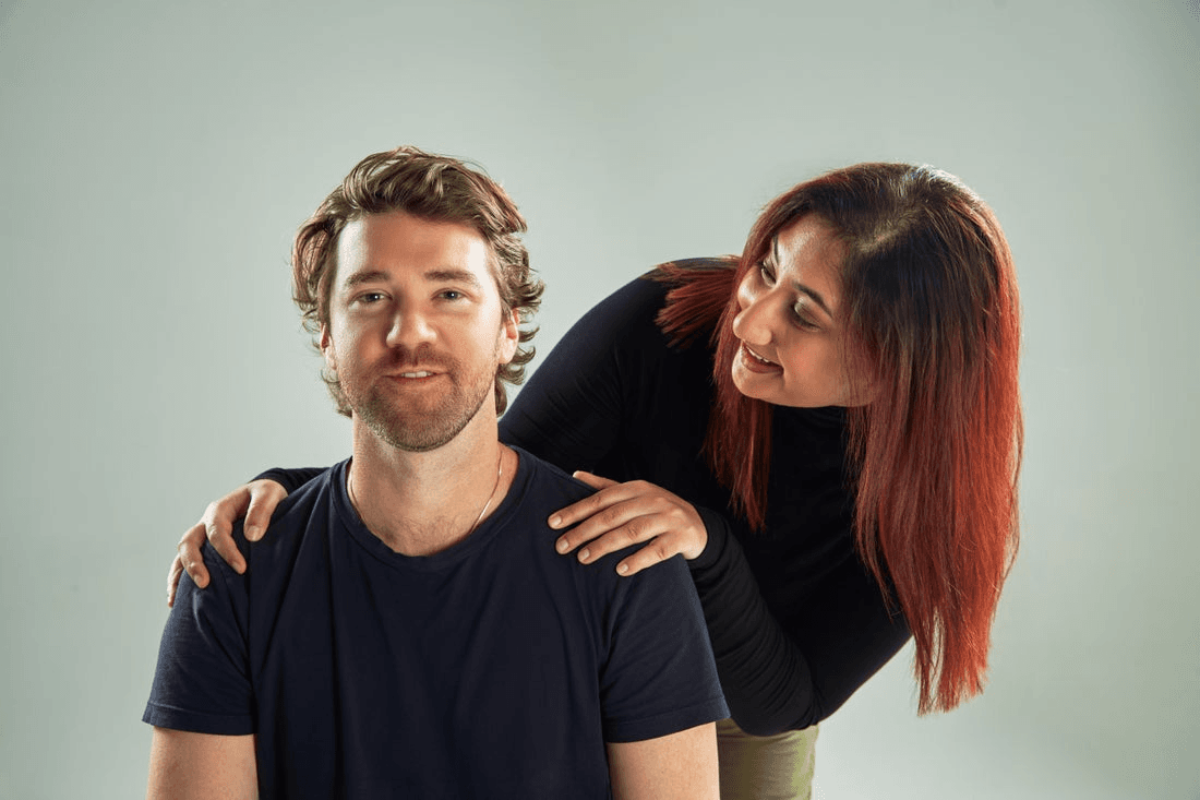 A sitting man looks forward while a woman, standing slightly behind him, smiles at him and holds his shoulders.